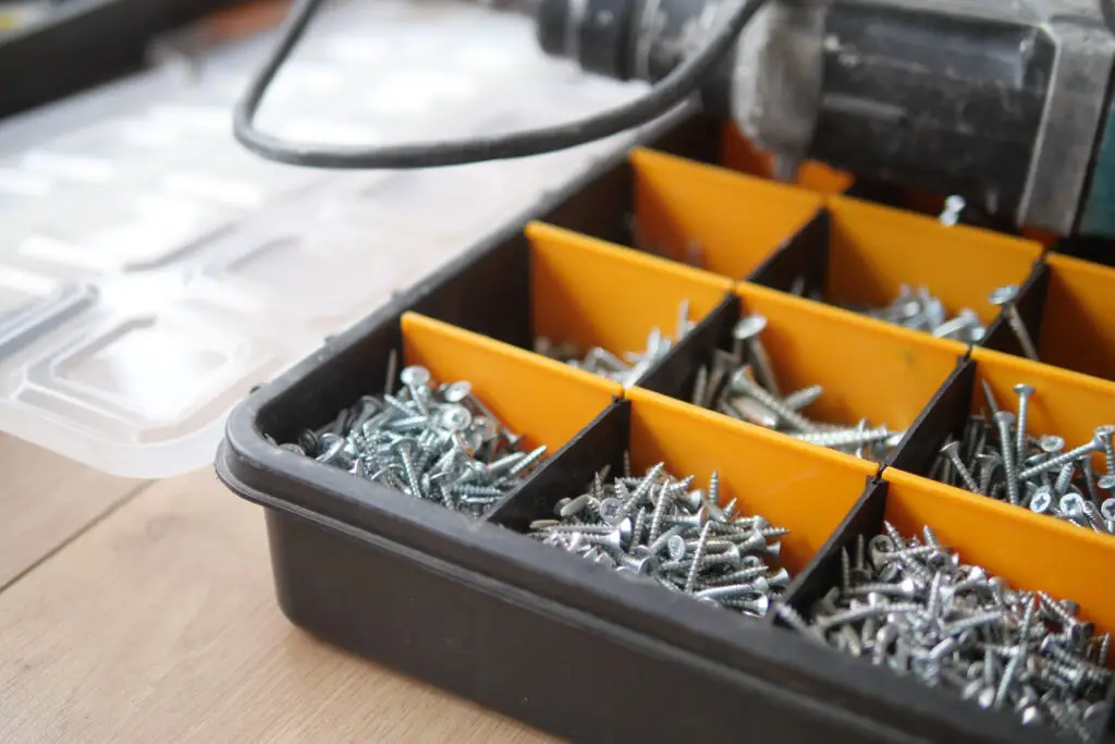 Caixa organizadora com divisórias laranja, cheia de parafusos de diferentes tamanhos e modelos. Ao fundo, uma tampa transparente e uma furadeira, sugerindo um ambiente de trabalho ou montagem. A imagem transmite organização e eficiência na escolha dos fixadores certos para cada necessidade.