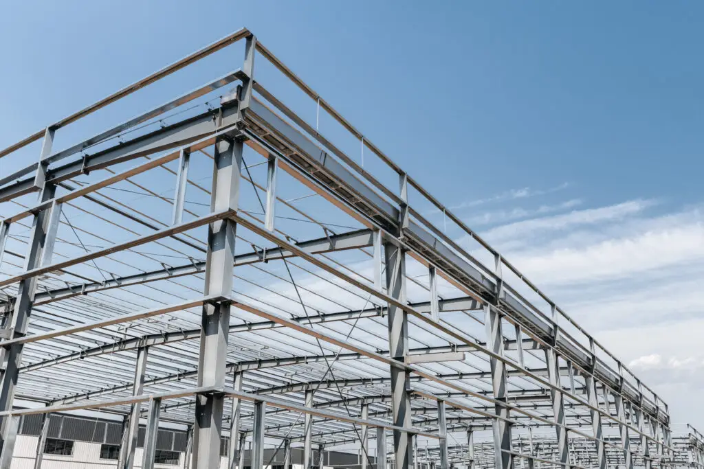 Estrutura metálica de grande porte em construção, composta por vigas e colunas de aço interligadas por parafusos de fixação, sob um céu azul claro.